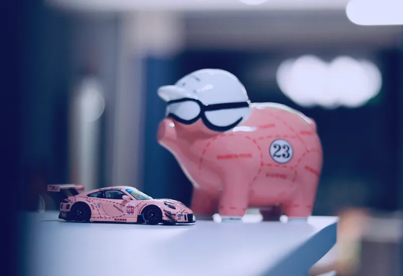 Pink Pig Porsche piggy bank wearing goggles and a helmet next to a matching pink race car model